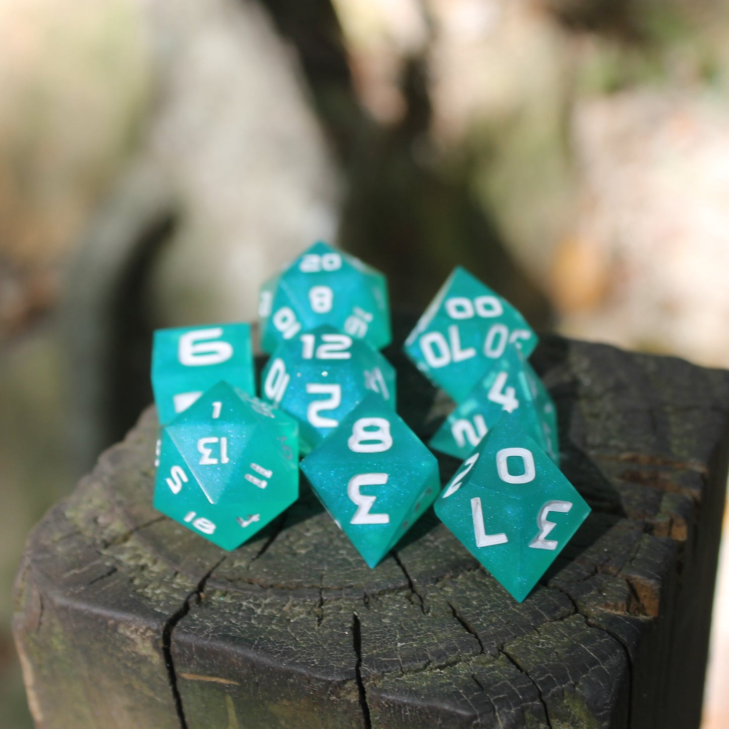 A set of polyhedral dnd dice that are teal with a pearlescent shine and have white numbers sits on top of a wooden post in direct sunlight.
