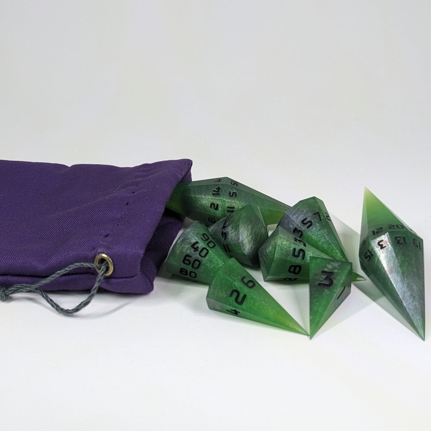 An 8 piece dagger shaped dice set that is venomous green with hints of silver like a poisoned blade with black numbers appear to spill out of a small, purple dice bag on a white background.