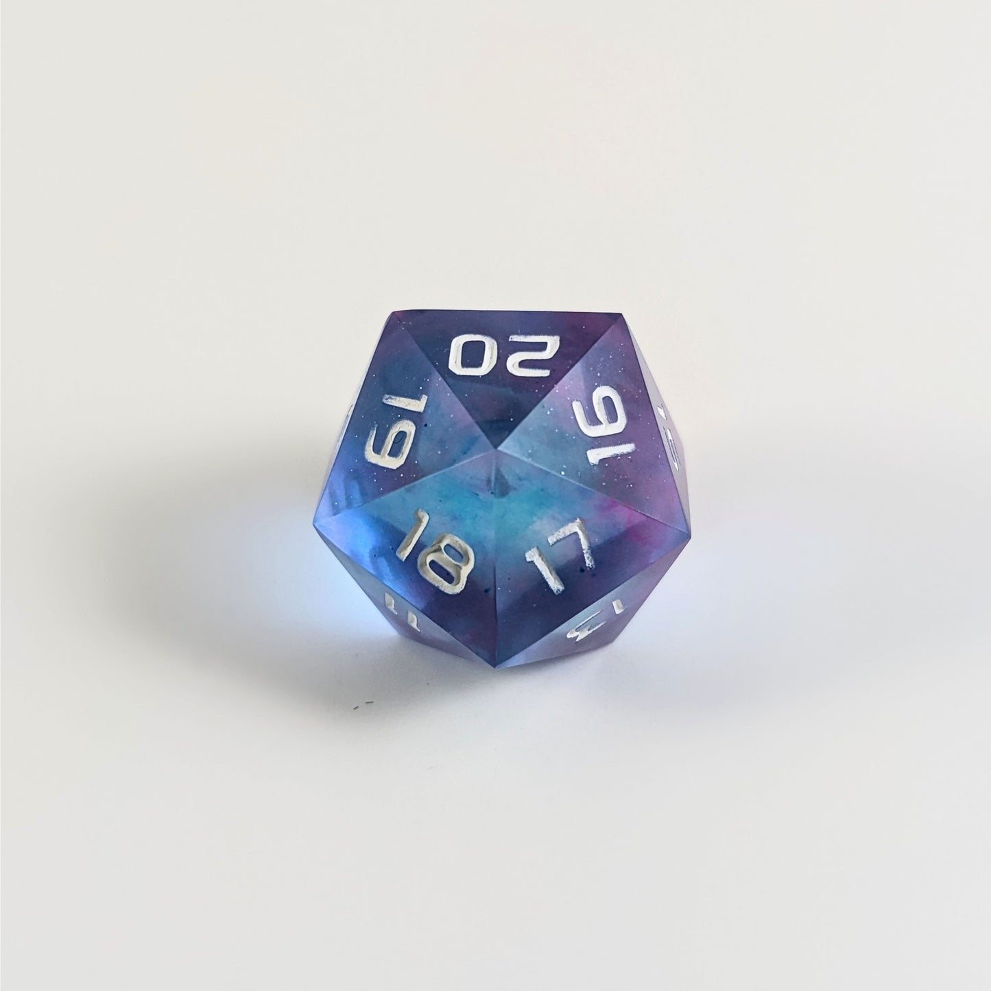 A low angle of a clear pink and teal spindown d20 with white numbers and a right spin sits on a white background with the 20 through 16 corner facing the camera showing the spiral of the spin down.