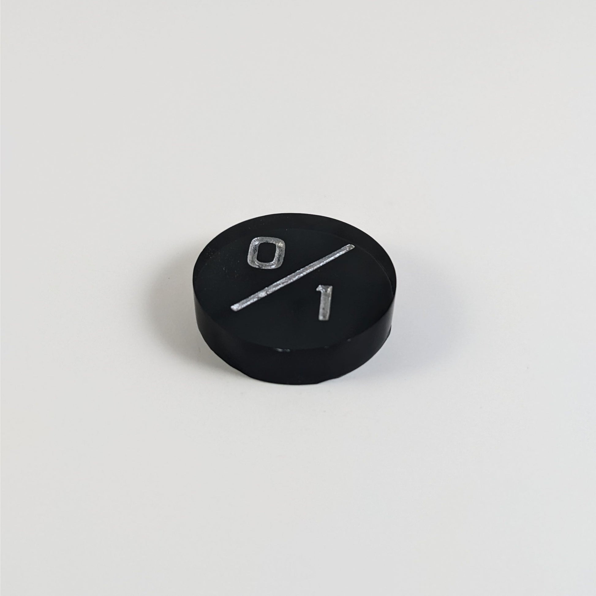 A low angle photo of a black 0/1 token with silver numbers sits on a white background.