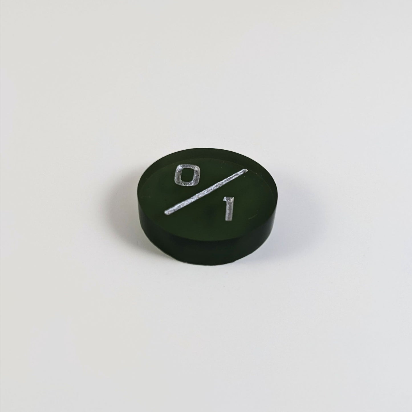 A low angle photo of a dark green 0/1 token with silver numbers sits on a white background.