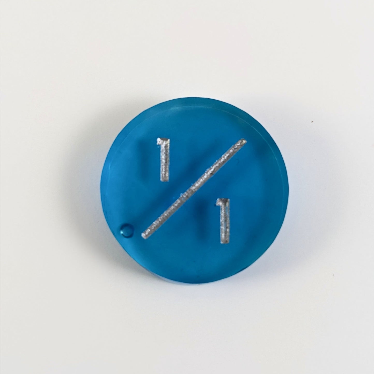 A top down photo of a blue 1/1 token with silver numbers sits on a white background. A bubble is visible near the bottom left of the slash but is clearly on the back of the token.