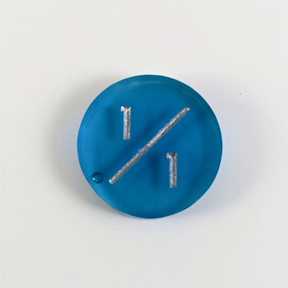 A top down photo of a blue 1/1 token with silver numbers sits on a white background. A bubble is visible near the bottom left of the slash but is clearly on the back of the token.