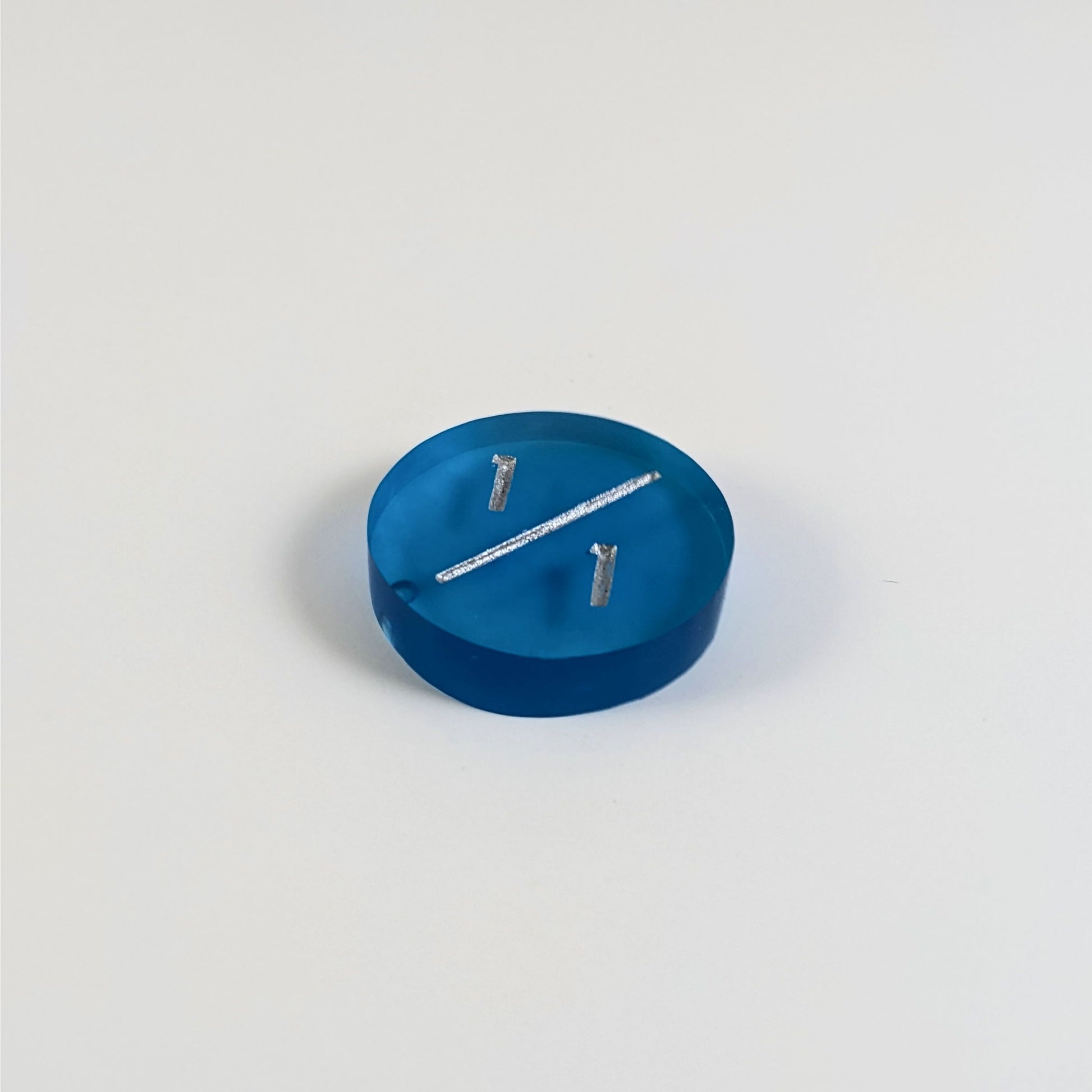 A low angle photo of a blue 1/1 token with silver numbers sits on a white background. The bubble is visible at the bottom end of the slash and is on the back of the token.