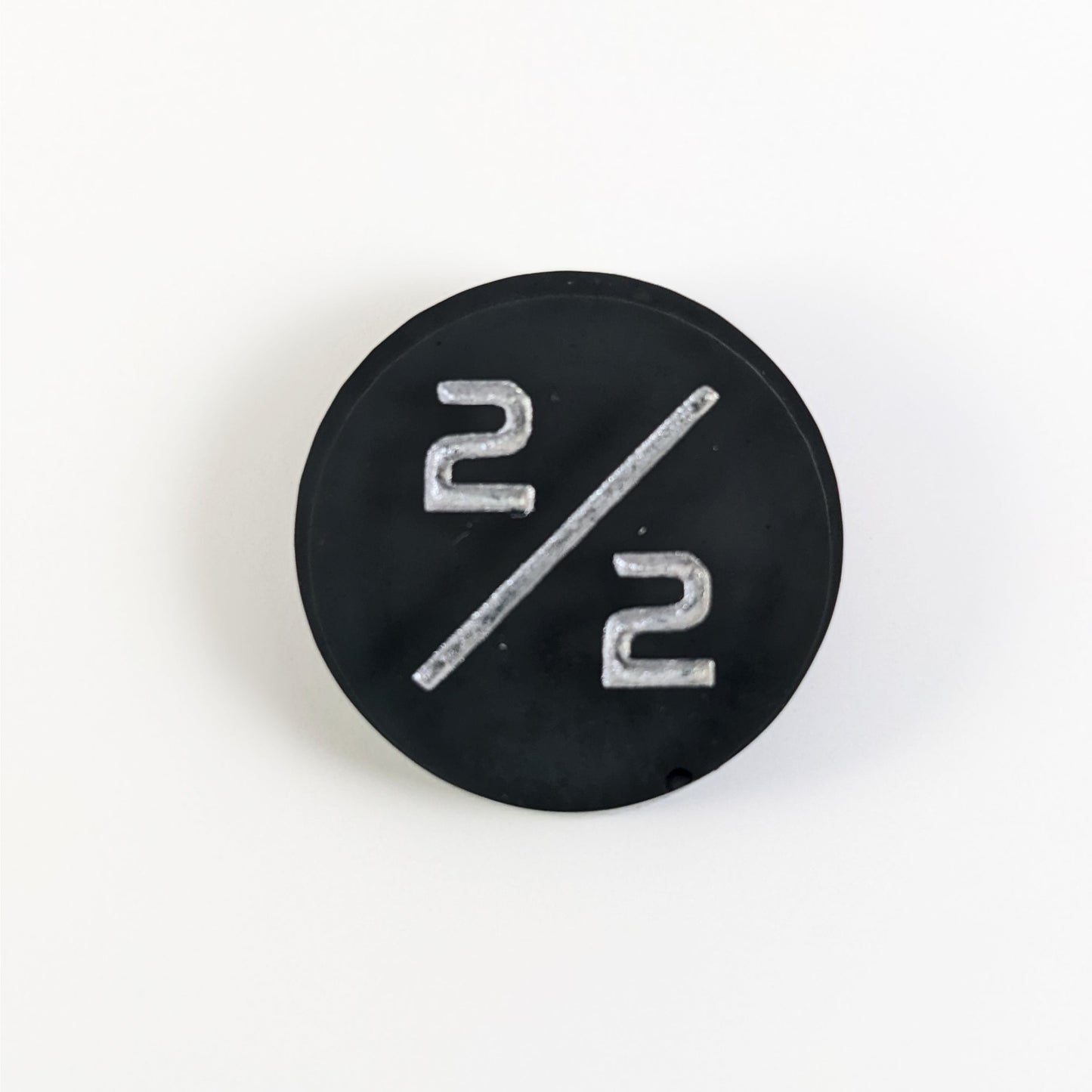 A top down photo of a black 2/2 token with silver numbers sits on a white background. A bubble is visible near the bottom right on the edge but it is on the back side of the token.