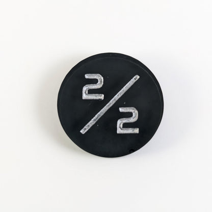 A top down photo of a black 2/2 token with silver numbers sits on a white background. A bubble is visible near the bottom right on the edge but it is on the back side of the token.