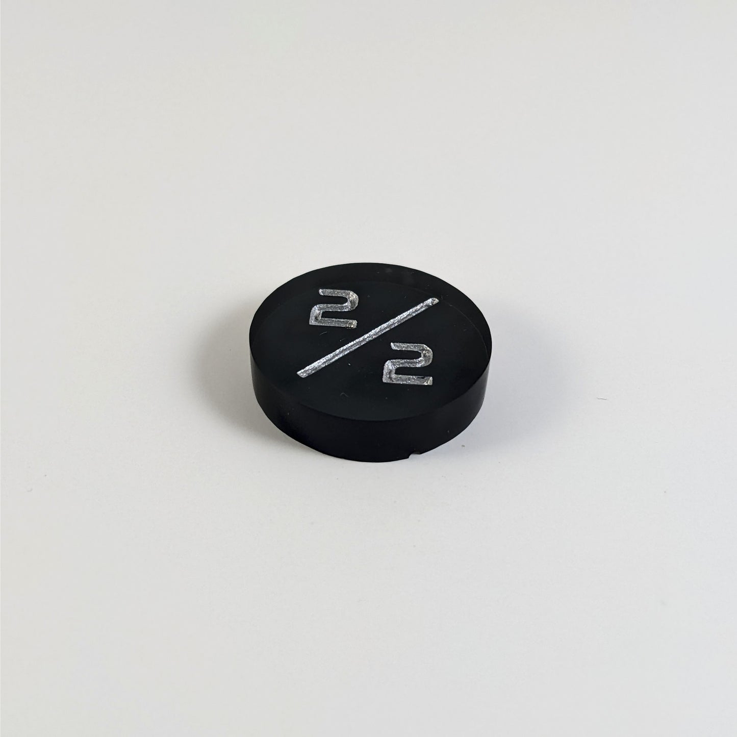 A low angle photo of a black 2/2 token with silver numbers sits on a white background. The small bubble is on the bottom edge of the token under the bottom 2.
