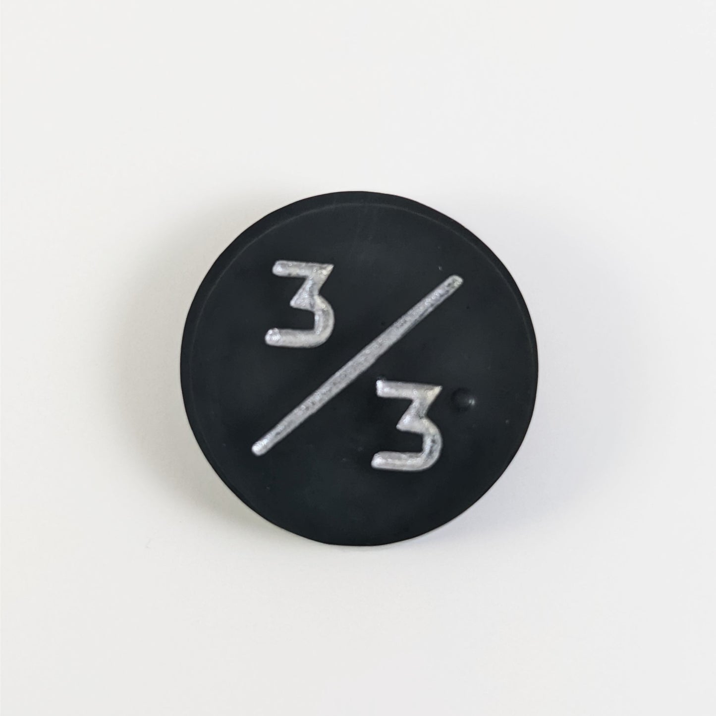 A top down photo of a black 3/3 token with silver numbers sits on a white background. A bubble is visible to the right of the bottom 3 but it is on the back side of the token.