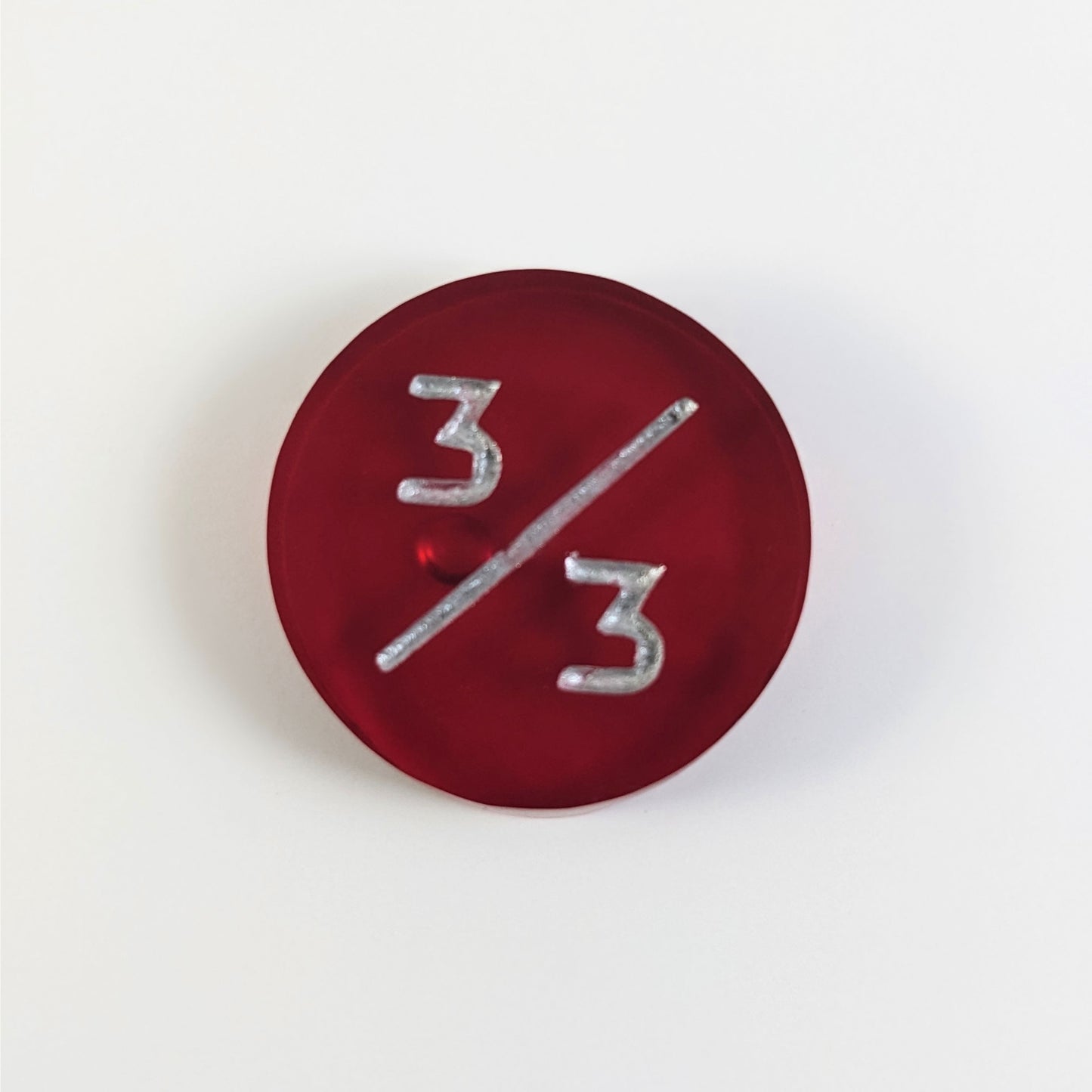 A top down photo of a red 3/3 token with silver numbers sits on a white background. A bubble is visible under the top 3 but it is on the back side of the token.