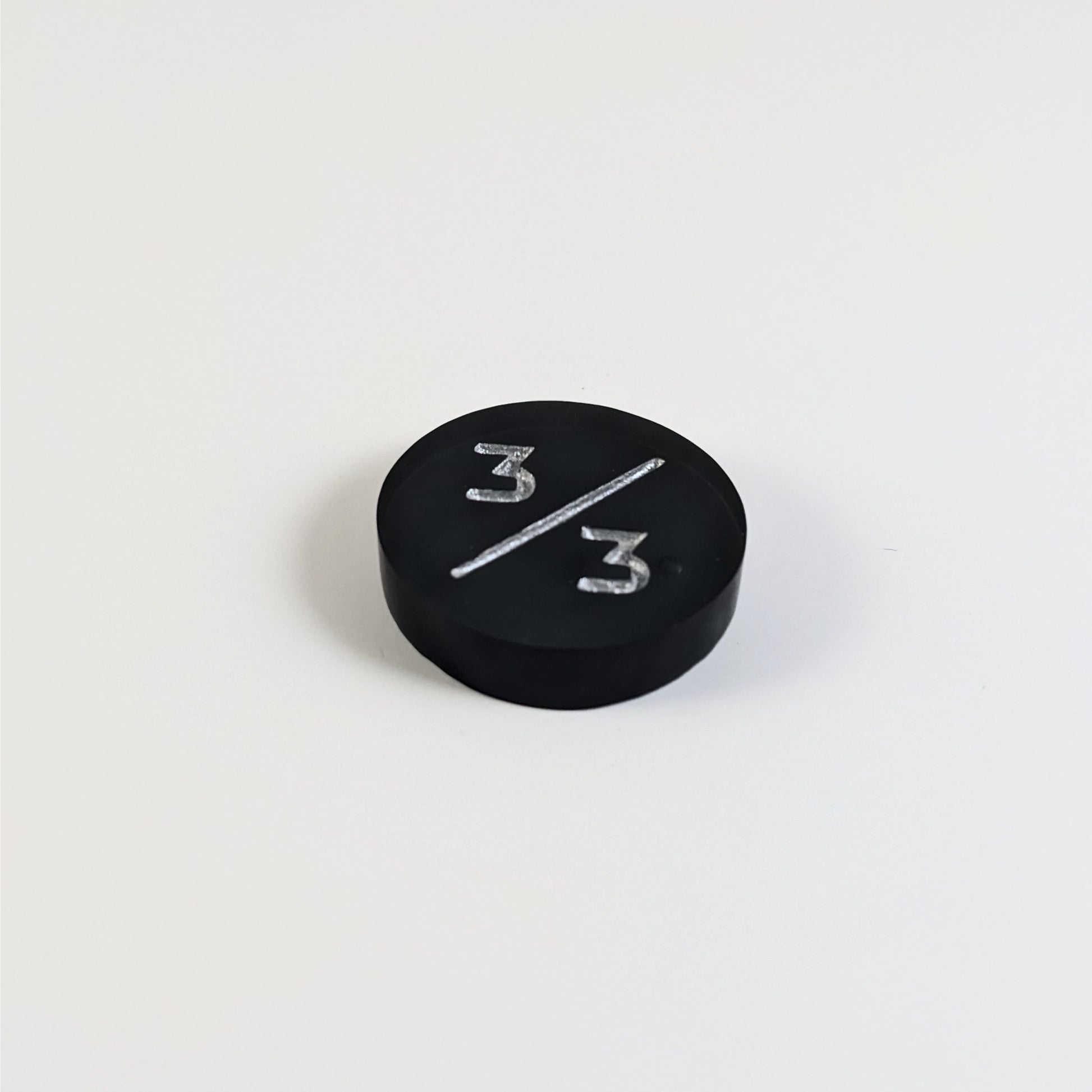 A low angle photo of a black 3/3 token with silver numbers sits on a white background. The bubble is visible to the right of the bottom 3 and is on the back of the token.