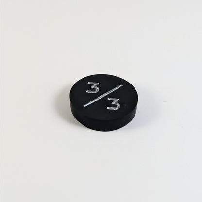 A low angle photo of a black 3/3 token with silver numbers sits on a white background. The bubble is visible to the right of the bottom 3 and is on the back of the token.