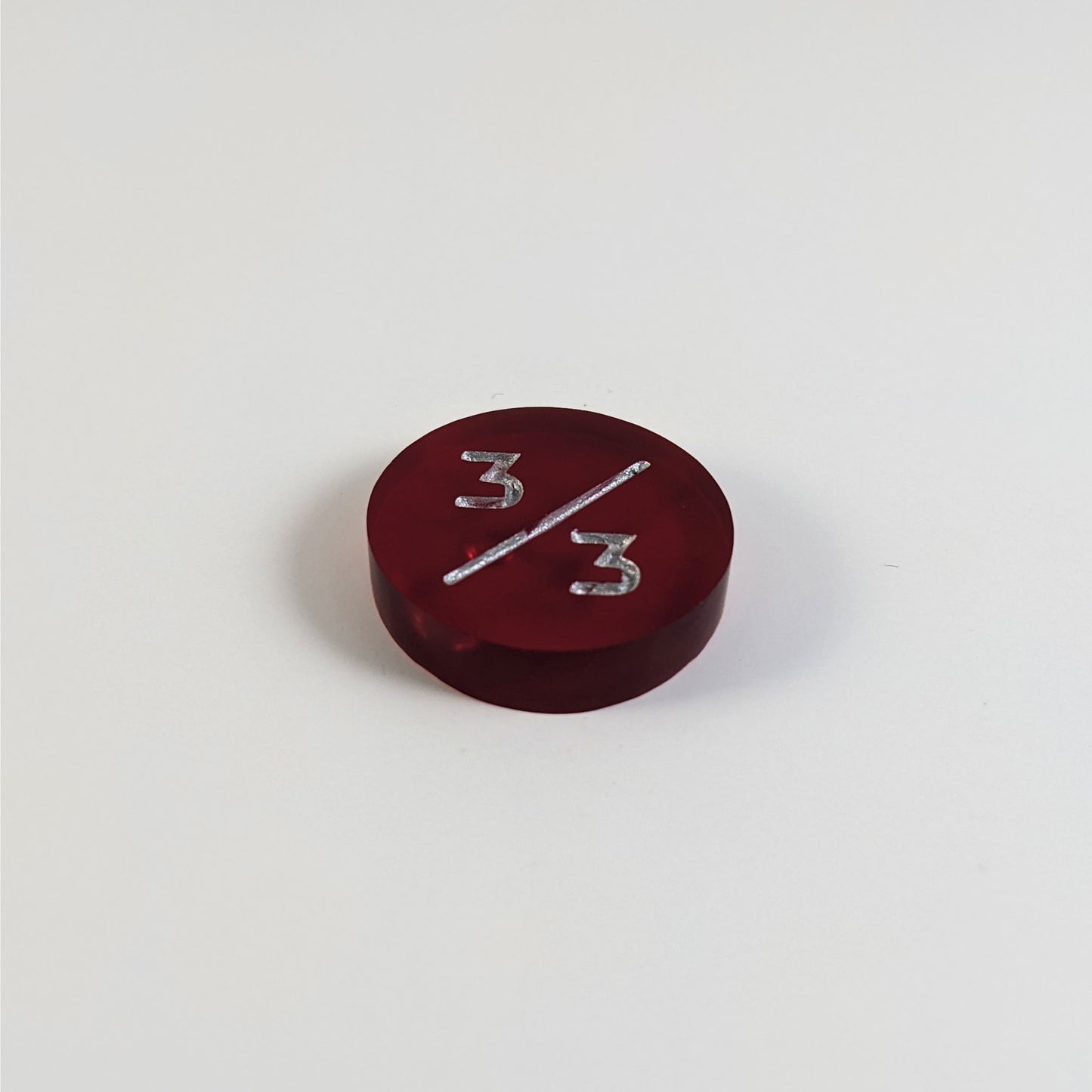 A low angle photo of a red 3/3 token with silver numbers sits on a white background. The bubble is visible under the top 3 but slightly masked by the slash as it is on the back side of the token.