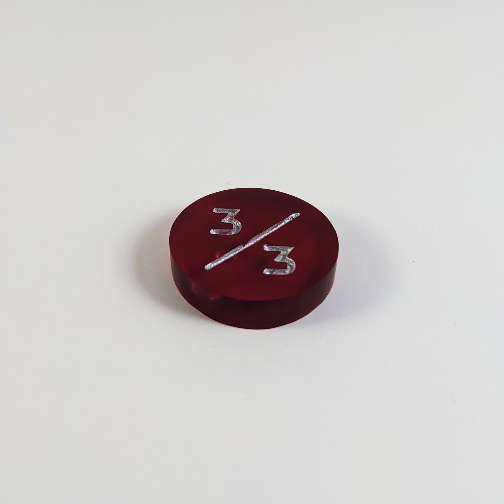 A low angle photo of a red 3/3 token with silver numbers sits on a white background. The bubble is visible under the top 3 but slightly masked by the slash as it is on the back side of the token.