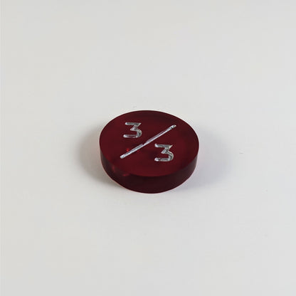 A low angle photo of a red 3/3 token with silver numbers sits on a white background. The bubble is visible under the top 3 but slightly masked by the slash as it is on the back side of the token.