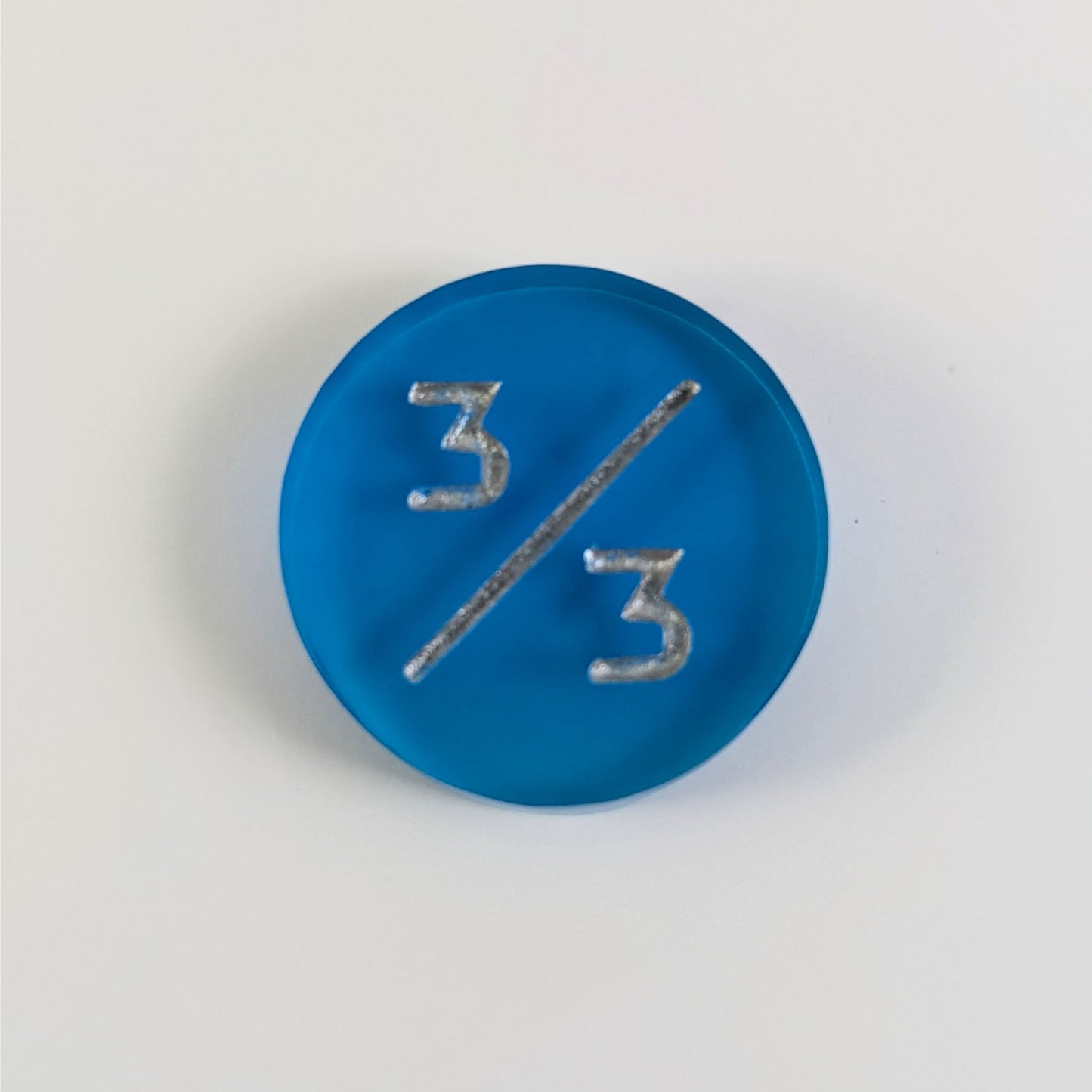 A top down photo of a blue 3/3 token with silver numbers sits on a white background.