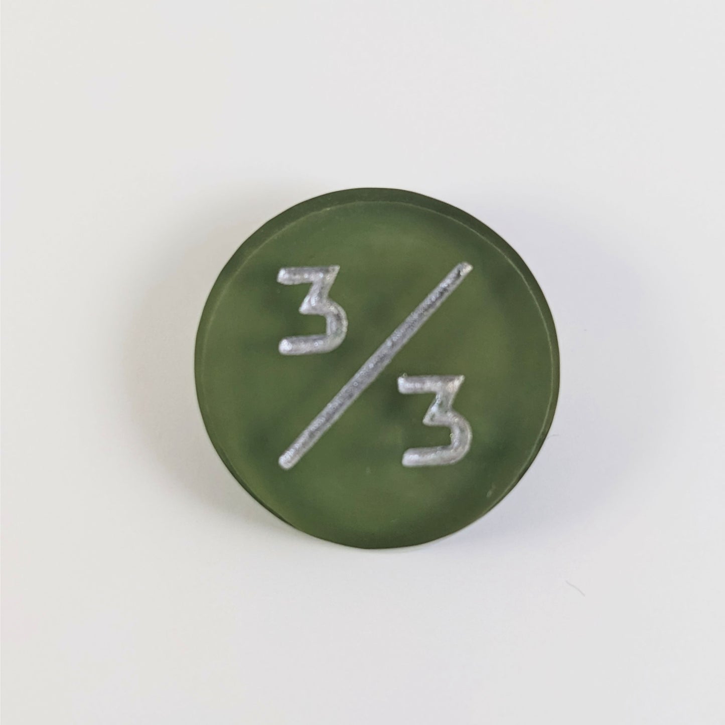 A top down photo of a dark green 3/3 token with silver numbers sits on a white background.