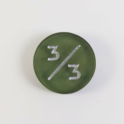 A top down photo of a dark green 3/3 token with silver numbers sits on a white background.