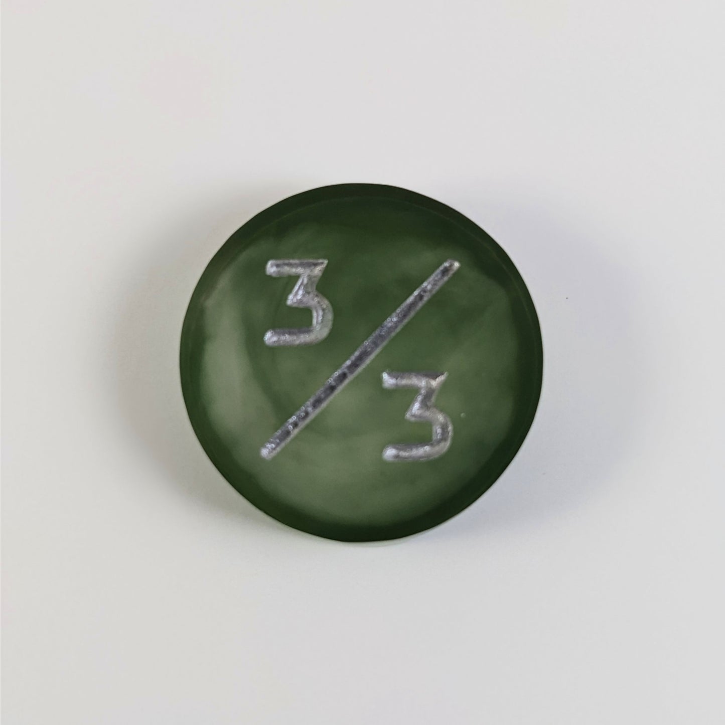 A top down photo of a dark green and white 3/3 token with silver numbers sits on a white background.