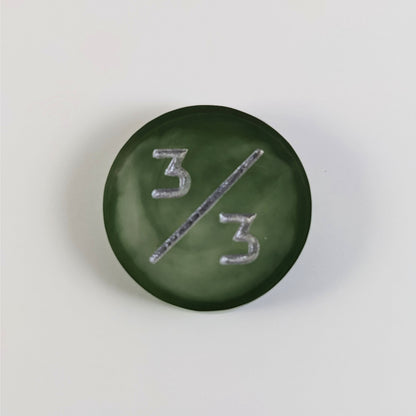 A top down photo of a dark green and white 3/3 token with silver numbers sits on a white background.