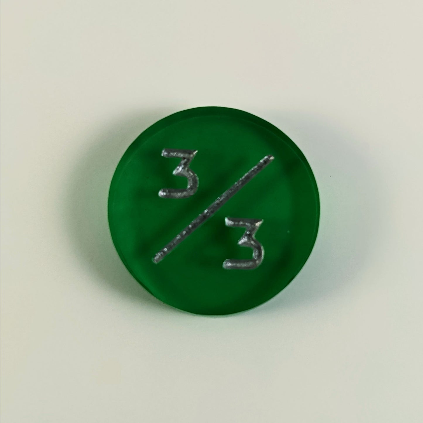 A top down photo of a green 3/3 token with silver numbers sits on a white background.