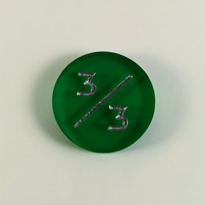 A top down photo of a green 3/3 token with silver numbers sits on a white background.