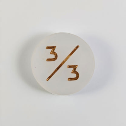 A top down photo of a white 3/3 token with gold numbers sits on a white background.