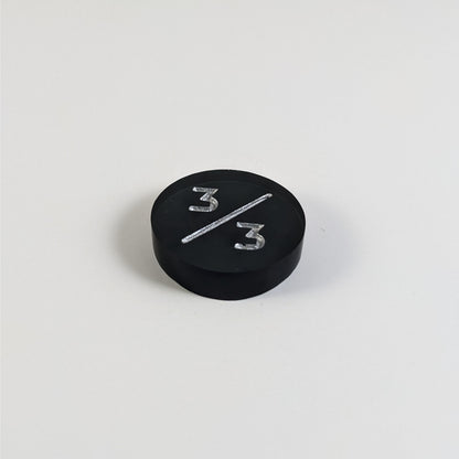 A low angle photo of a black 3/3 token with silver numbers sits on a white background.