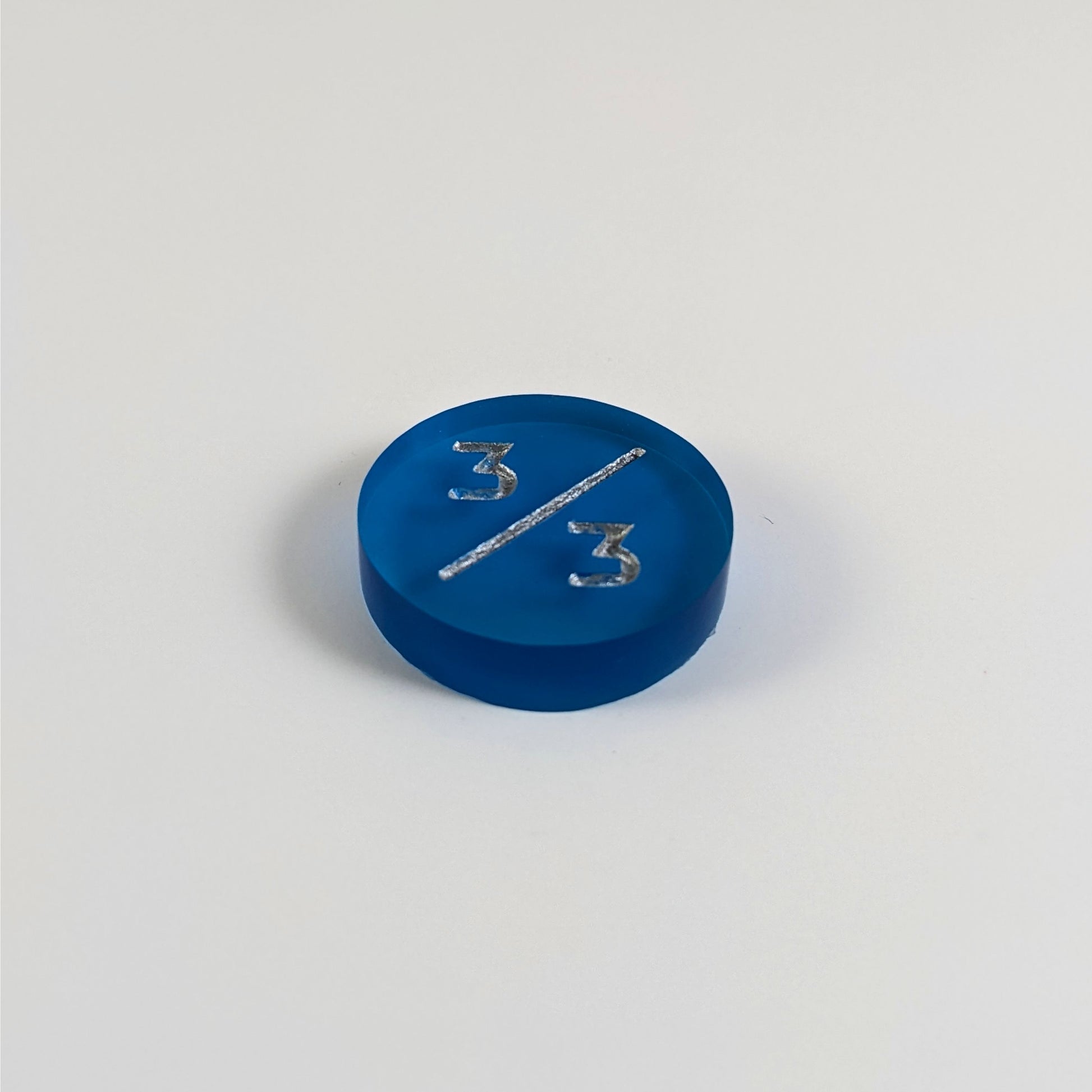 A low angle photo of a blue 3/3 token with silver numbers sits on a white background.