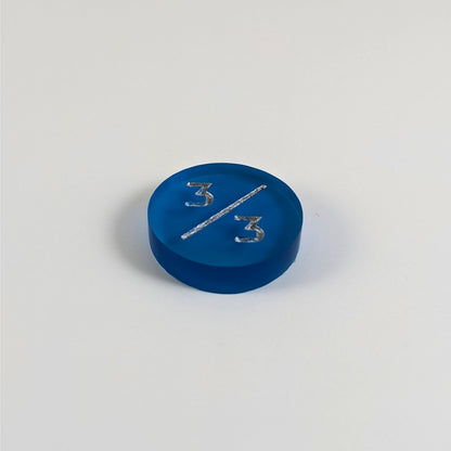 A low angle photo of a blue 3/3 token with silver numbers sits on a white background.
