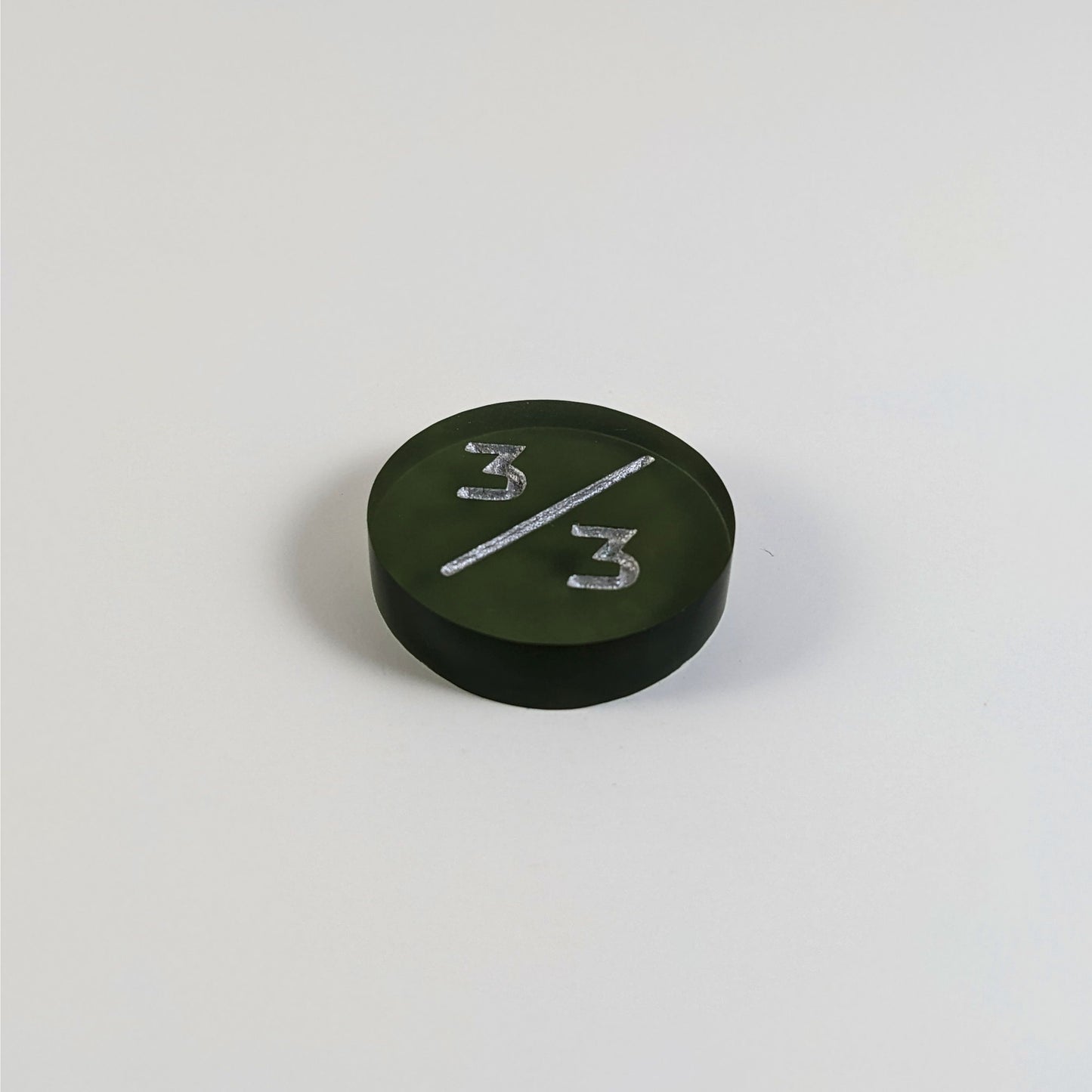 A low angle photo of a dark green 3/3 token with silver numbers sits on a white background.
