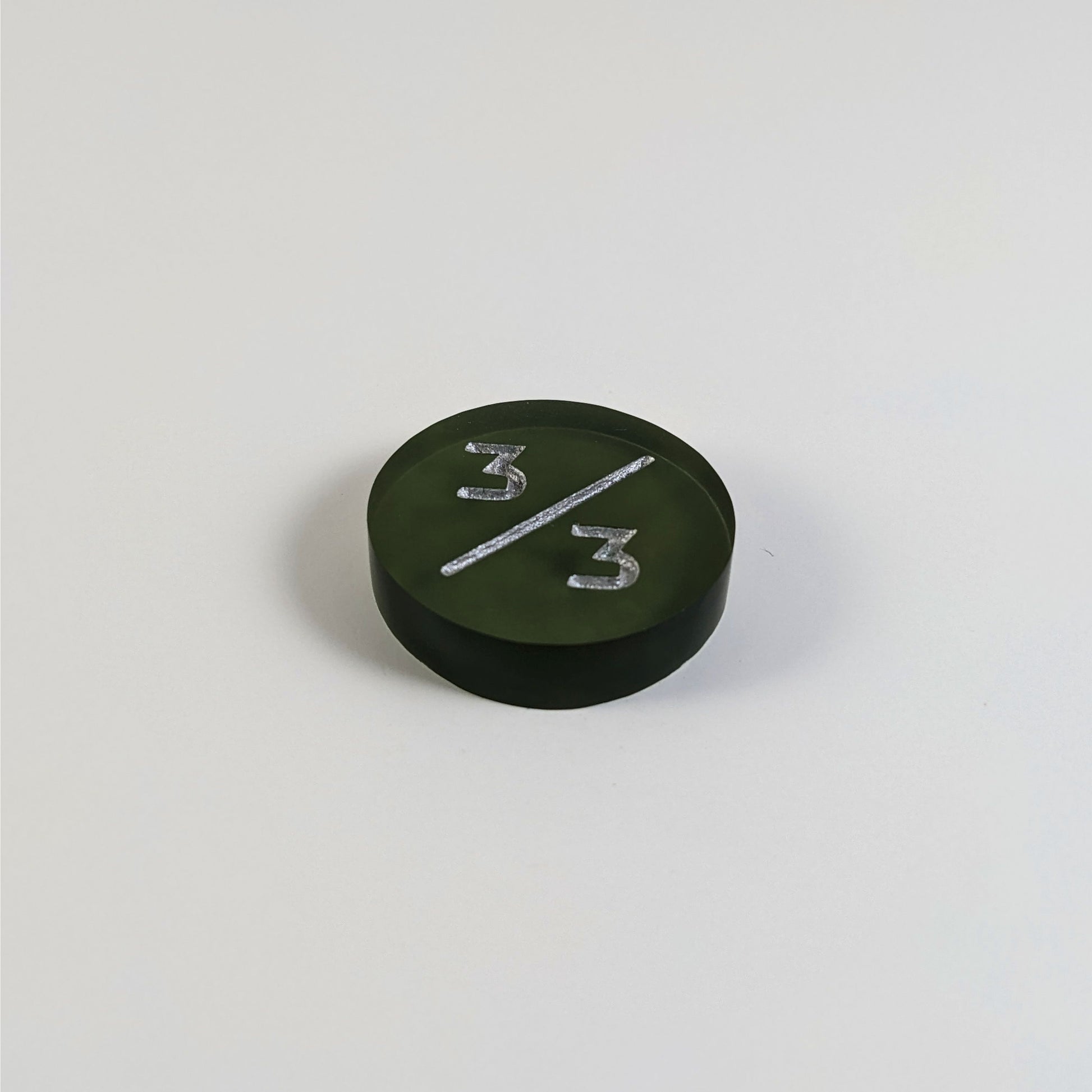 A low angle photo of a dark green 3/3 token with silver numbers sits on a white background.