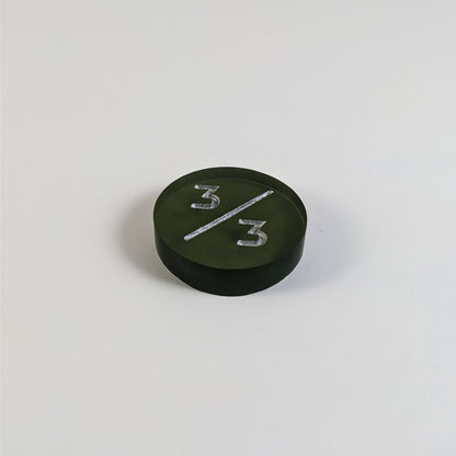 A low angle photo of a dark green 3/3 token with silver numbers sits on a white background.