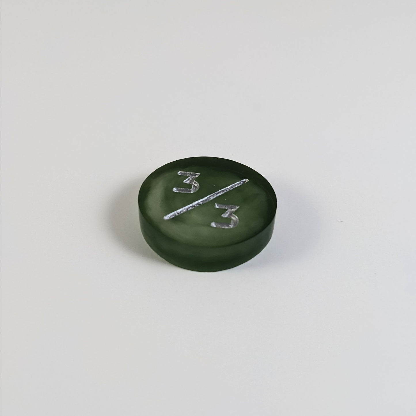 A low angle photo of a dark green and white 3/3 token with silver numbers sits on a white background.