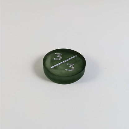 A low angle photo of a dark green and white 3/3 token with silver numbers sits on a white background.