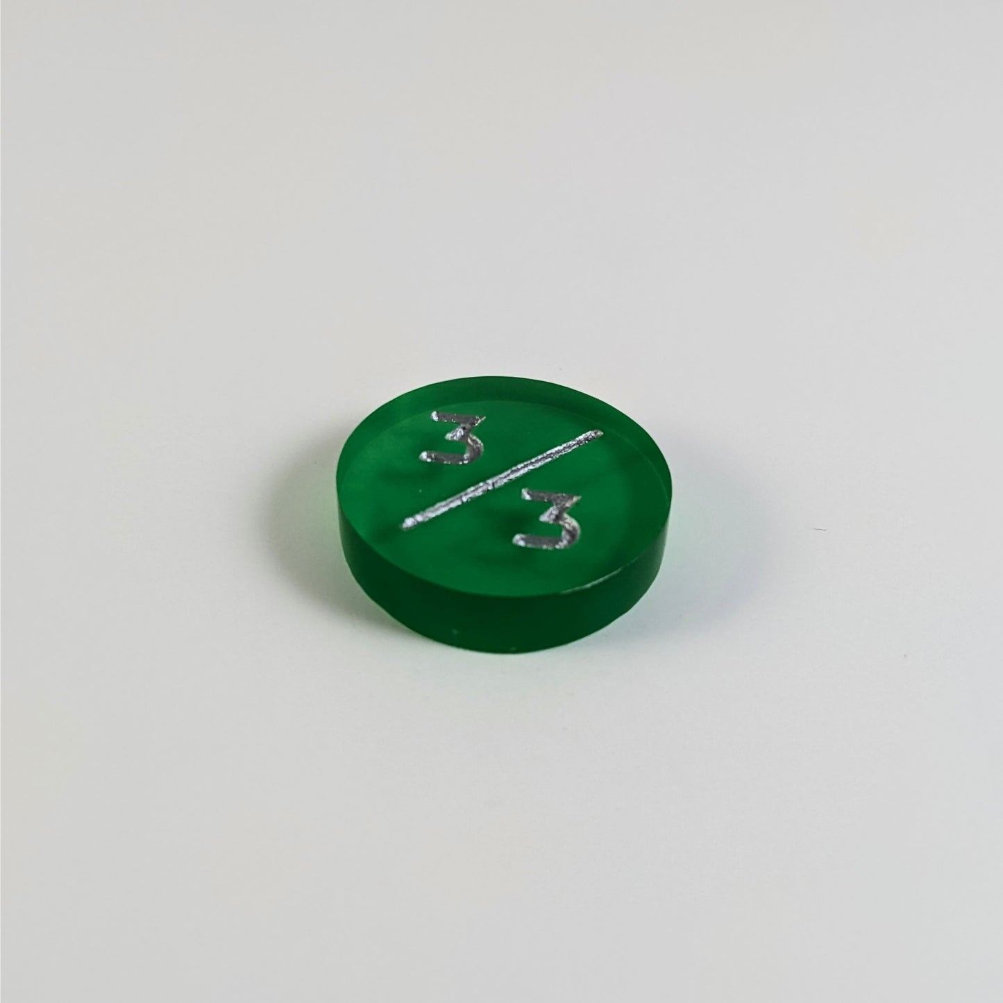 A low angle photo of a green 3/3 token with silver numbers sits on a white background.