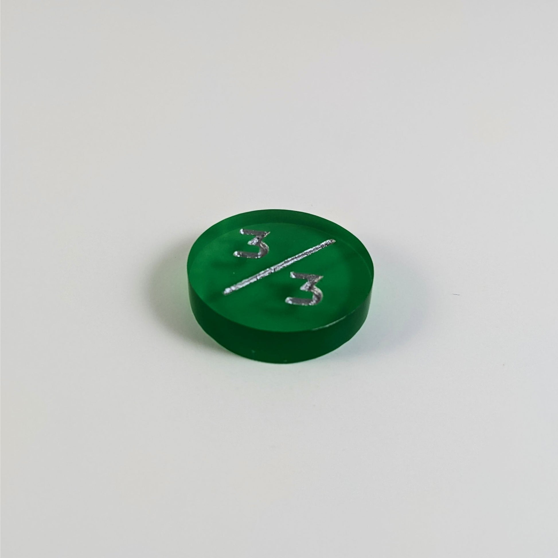 A low angle photo of a green 3/3 token with silver numbers sits on a white background.