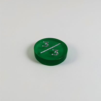 A low angle photo of a green 3/3 token with silver numbers sits on a white background.