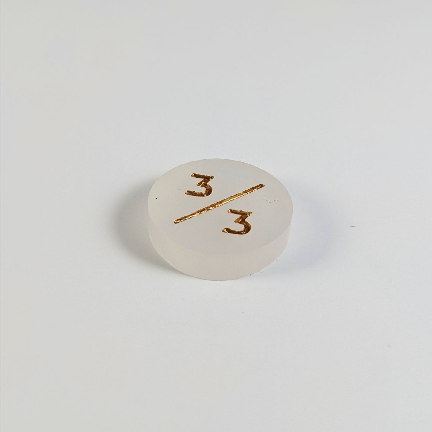 A low angle photo of a white 3/3 token with gold numbers sits on a white background.