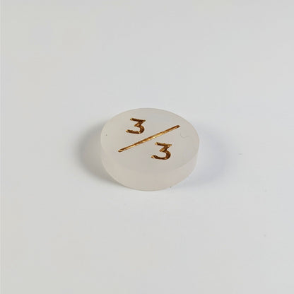 A low angle photo of a white 3/3 token with gold numbers sits on a white background.