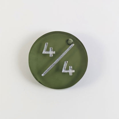 A top down photo of a dark green 4/4 token with silver numbers sits on a white background. A bubble is visible at the top right by the end of the slash, but it is on the back side of the token.