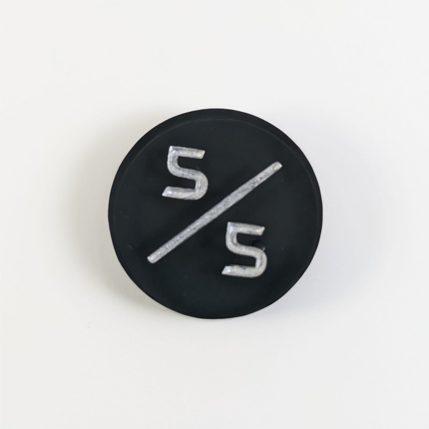 A top down photo of a black 5/5 token with silver numbers sits on a white background. A bubble is visible just behind the bottom 5, but it is on the back side of the token.