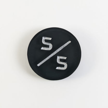 A top down photo of a black 5/5 token with silver numbers sits on a white background. A bubble is visible just behind the bottom 5, but it is on the back side of the token.