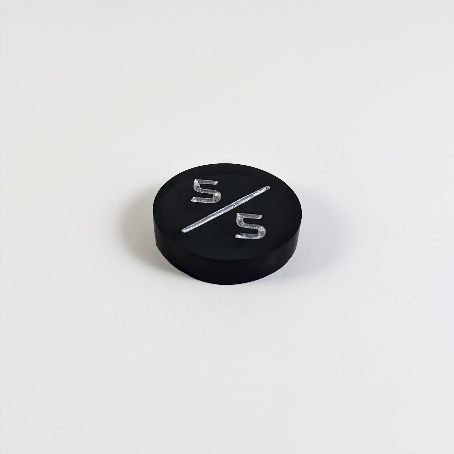 A low angle photo of a black 5/5 token with silver numbers sits on a white background. The cosmetic flaw is not apparently visible at this angle.