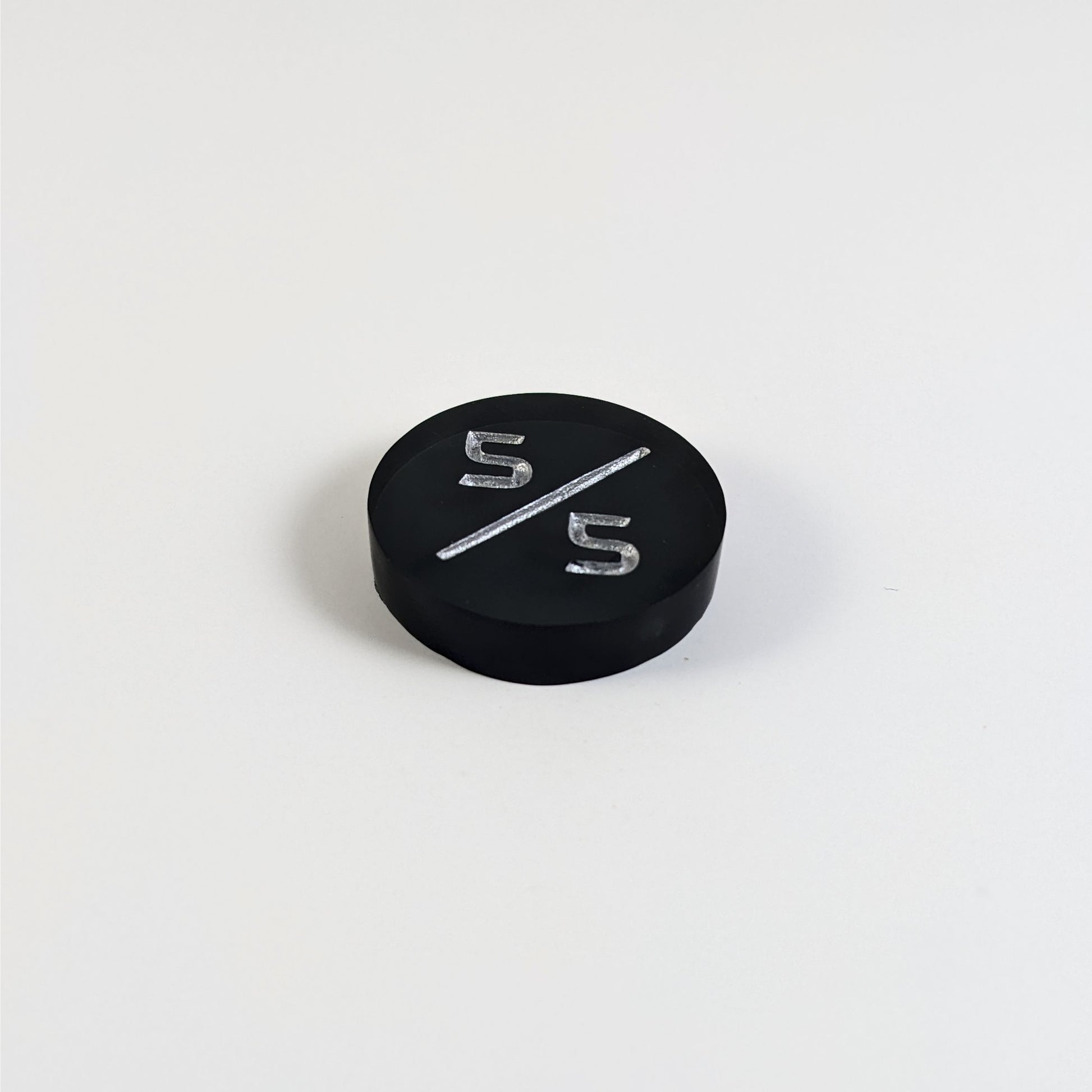 A low angle photo of a black 5/5 token with silver numbers sits on a white background. The cosmetic flaw is not apparently visible at this angle.