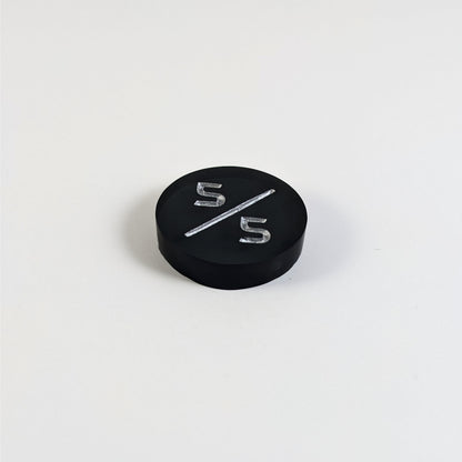 A low angle photo of a black 5/5 token with silver numbers sits on a white background. The cosmetic flaw is not apparently visible at this angle.