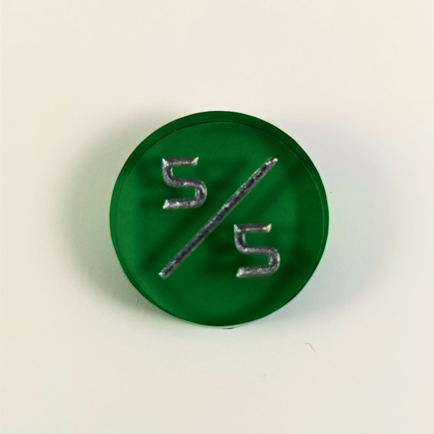 A top down photo of a green 5/5 token with silver numbers sits on a white background.