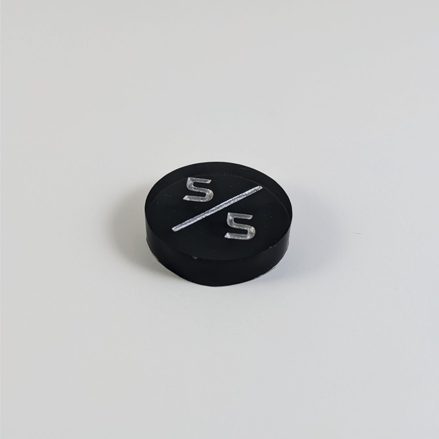 A low angle photo of a black 5/5 token with silver numbers sits on a white background.