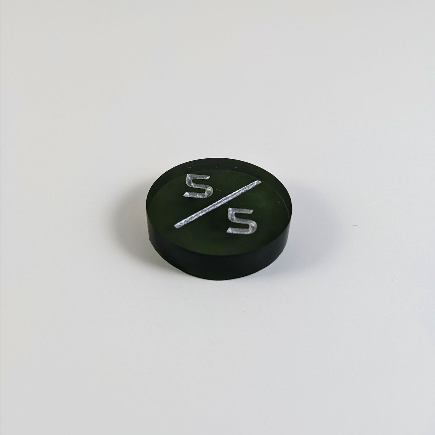 A low angle photo of a dark green 5/5 token with silver numbers sits on a white background.
