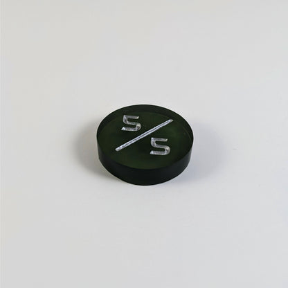 A low angle photo of a dark green 5/5 token with silver numbers sits on a white background.