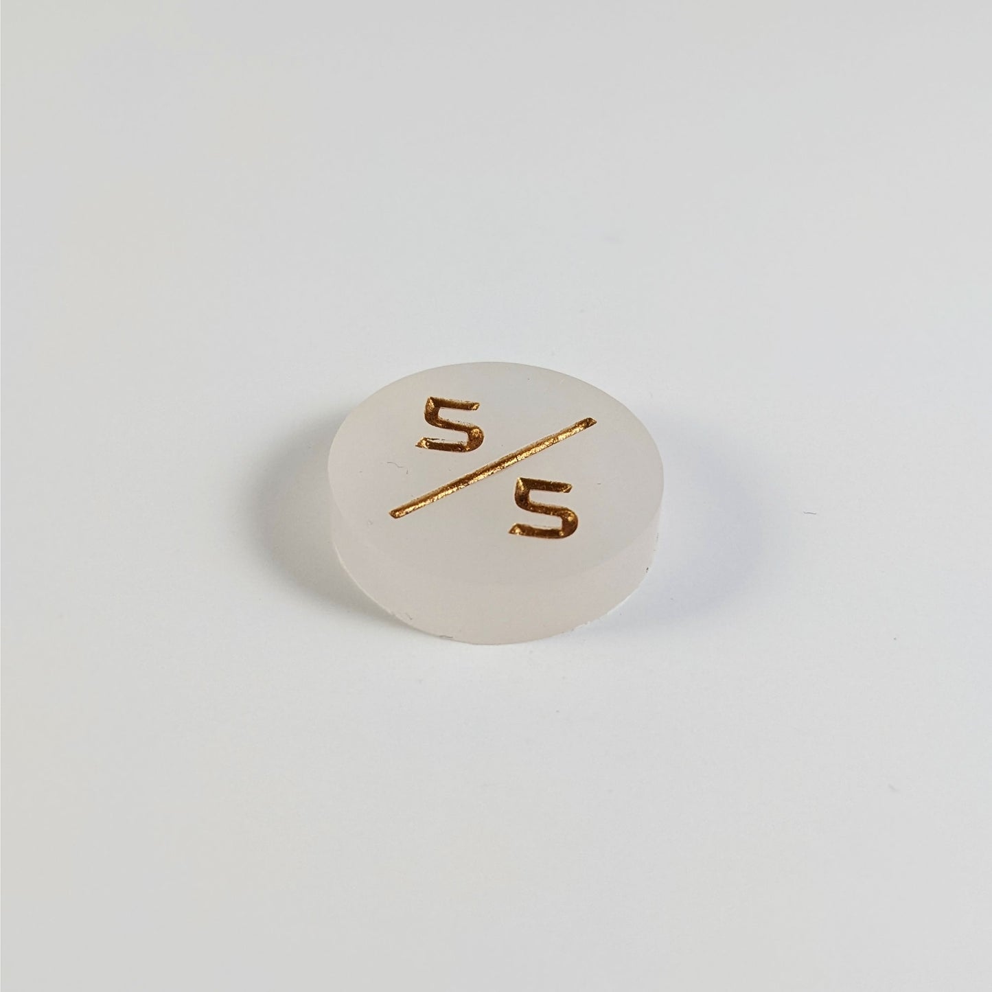 A low angle photo of a white 5/5 token with gold numbers sits on a white background.